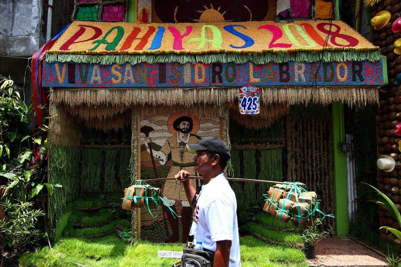 A man with bundles of food walking past a house decorated for the Pahiyas Festival