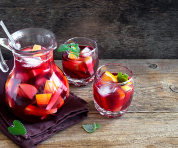 Picture shows a jug of red wine filled with chopped fruit alongside two glasses on top of a wooden table