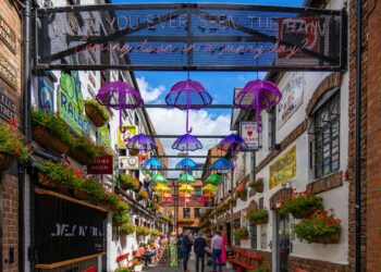 Picture showing Belfast's Cathedral Quarter. The street is lined with colourful umbrellas above and shows several pubs on either side of the street