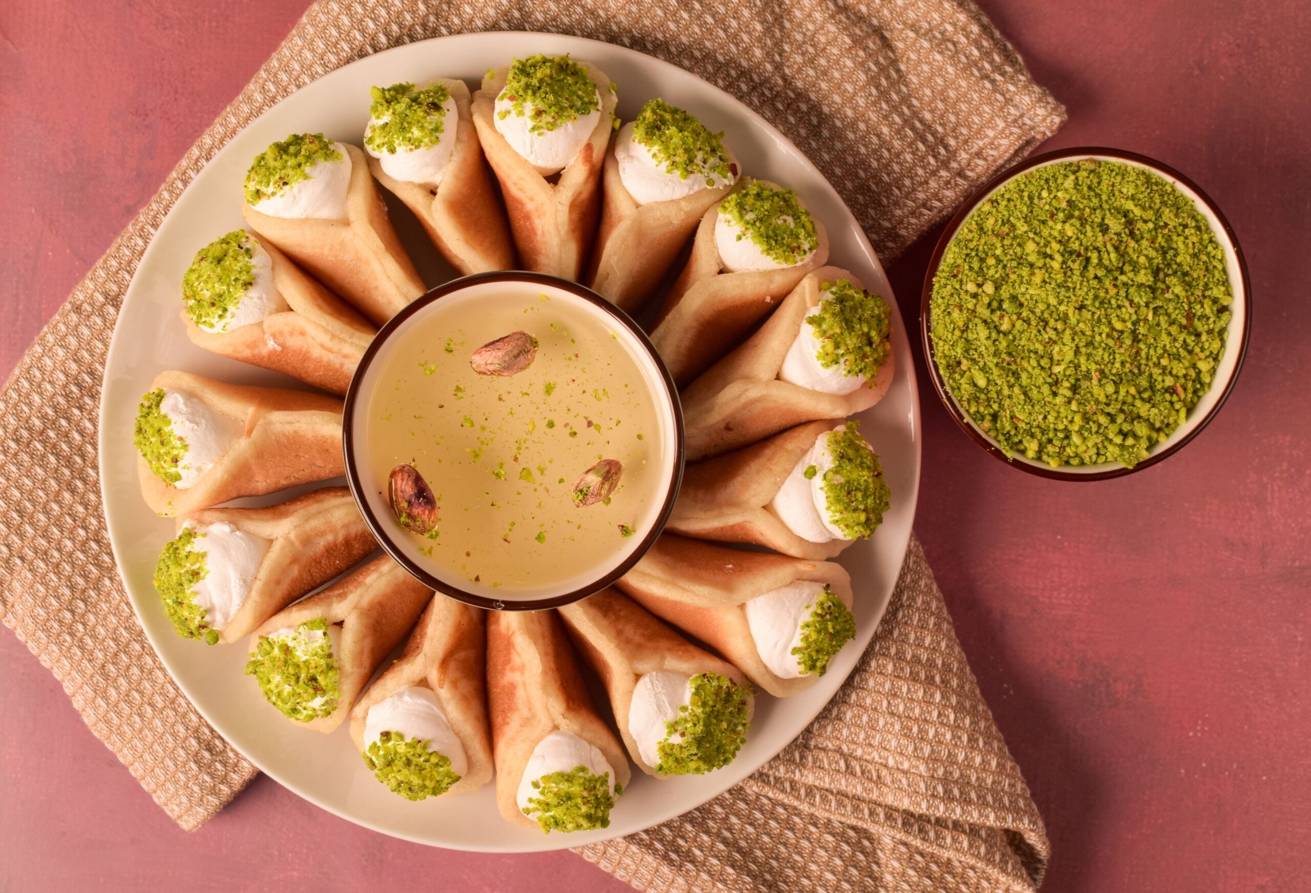 Mini pancake-like dessert stuffed with different fillings like cheese, custard, clotted cream and crushed nuts, and garnished with pistachio placed around a dip on a plate