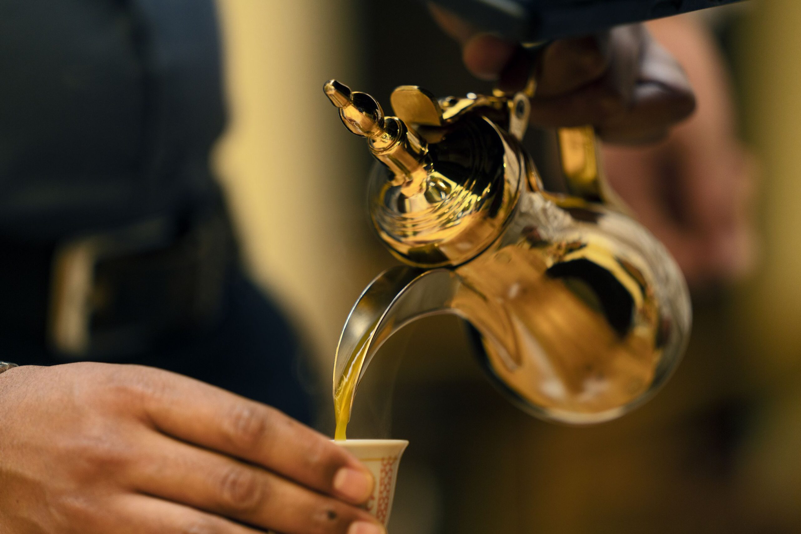 A pair of hand pouring a cup of coffee fro a gold metal teapot