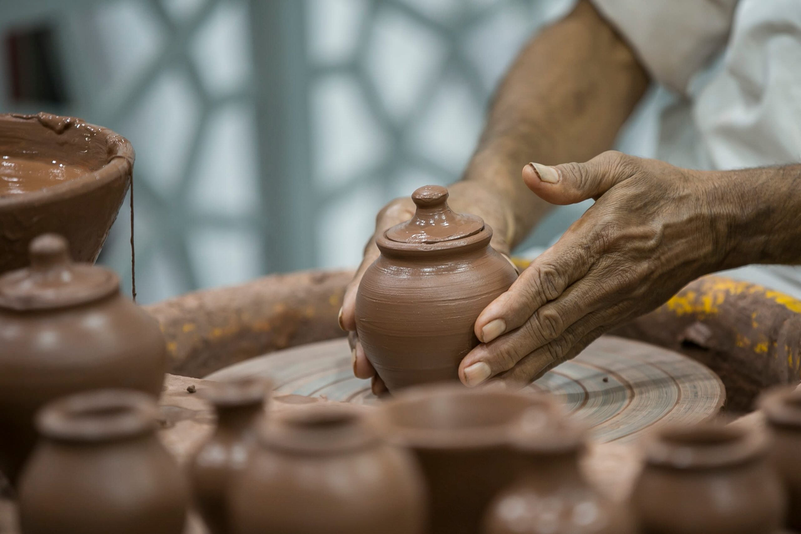 A pair of hands shaping a pottery jar with a lid on top on the spinning wheel