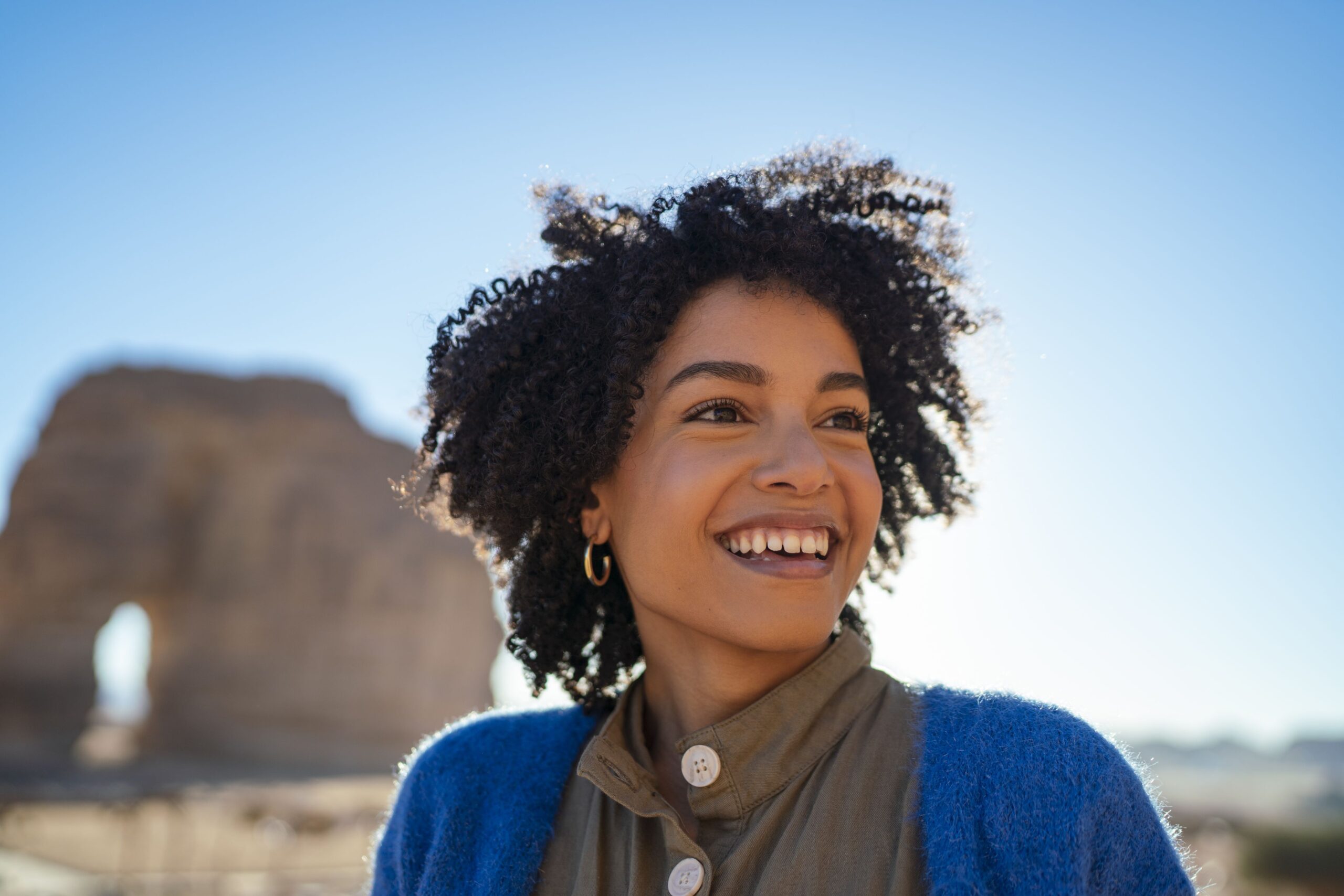 A lady in a blue cardigan stands smiling in the sunny desert